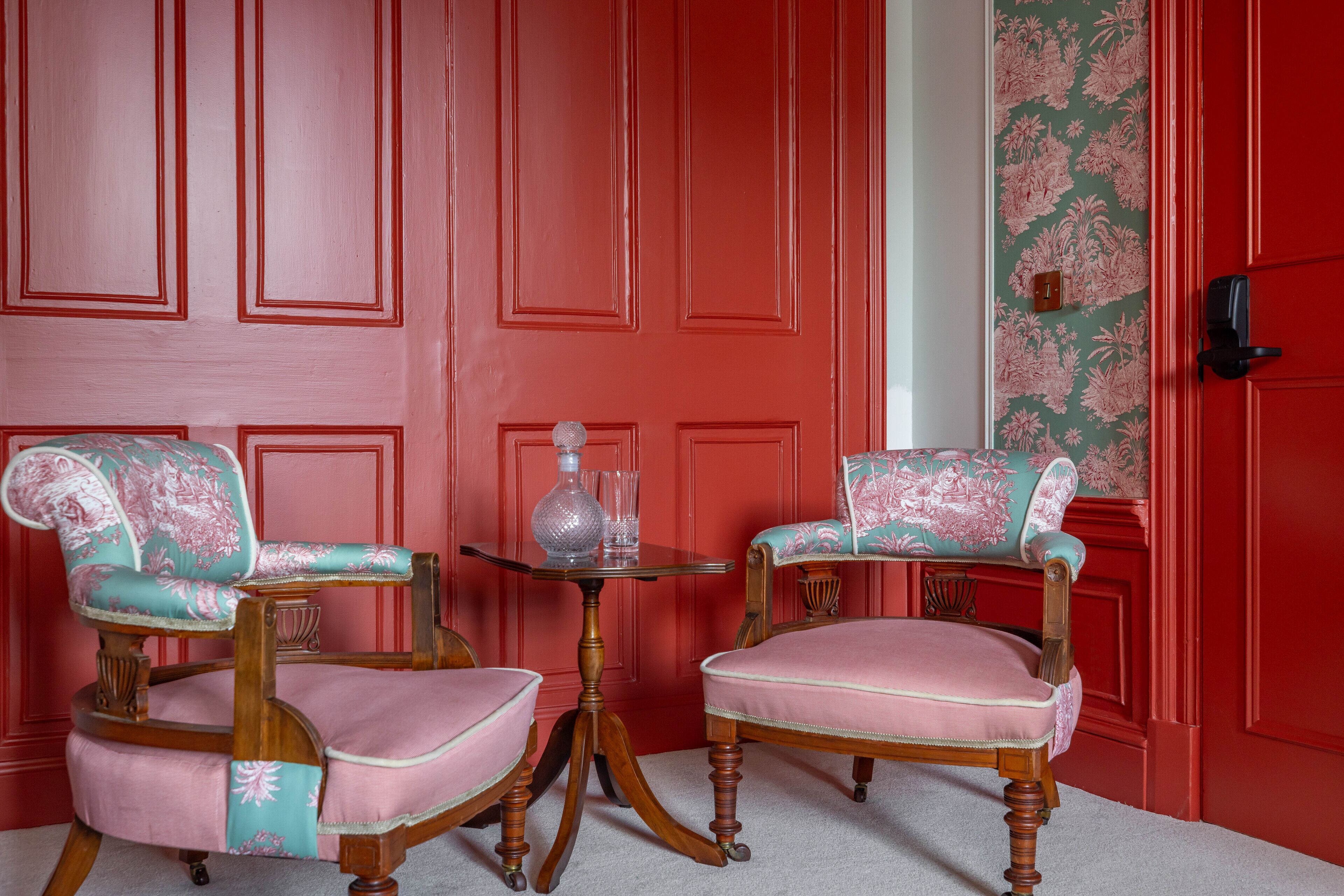 Dos sillones tapizados con motivos florales, un decantador de vidrio y una mesita frente a una puerta roja con paneles.