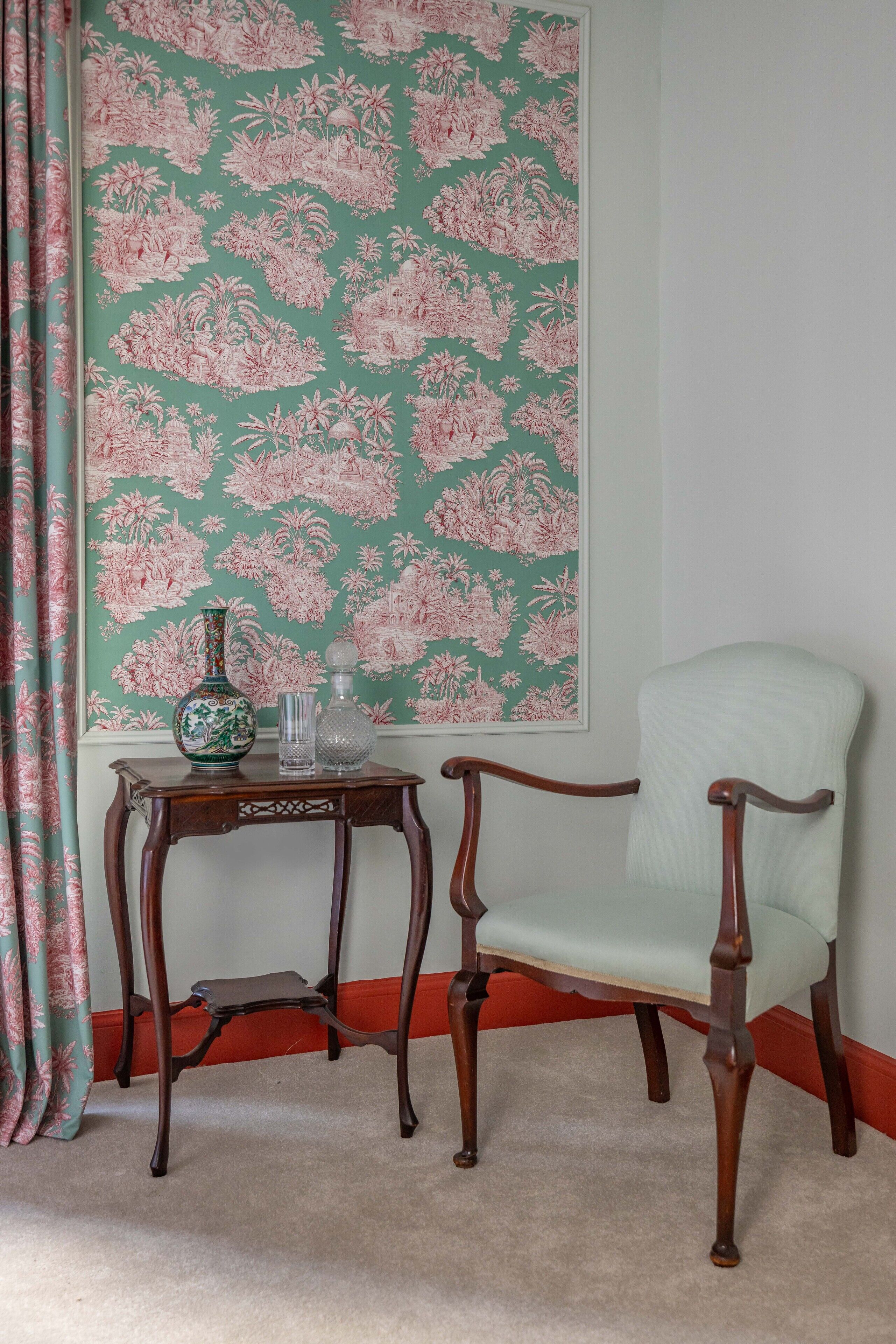 Une pièce avec un papier peint à motifs floraux, une chaise en bois, une petite table sur laquelle se trouvent un vase et un verre, et un plintheur rouge.