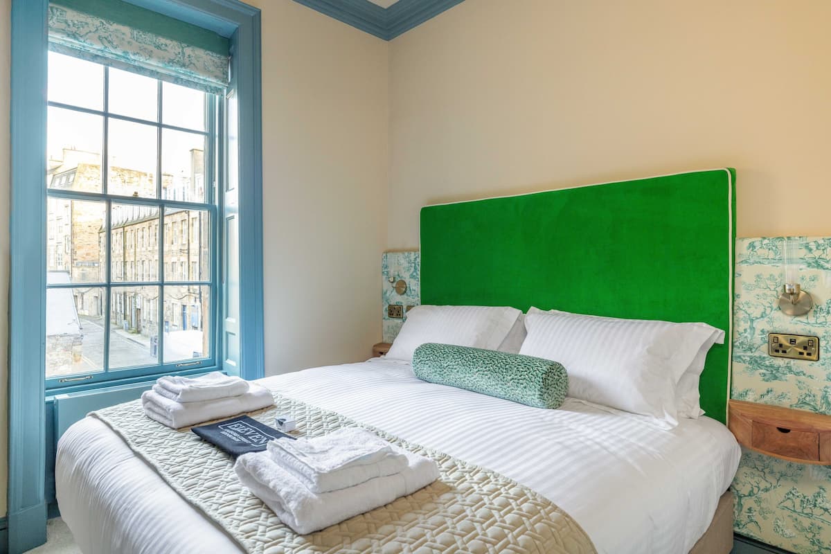 Een keurig opgemaakt bed met een groen hoofdeinde, een raam met blauwe omlijsting en een muur met bloemenbehang.
