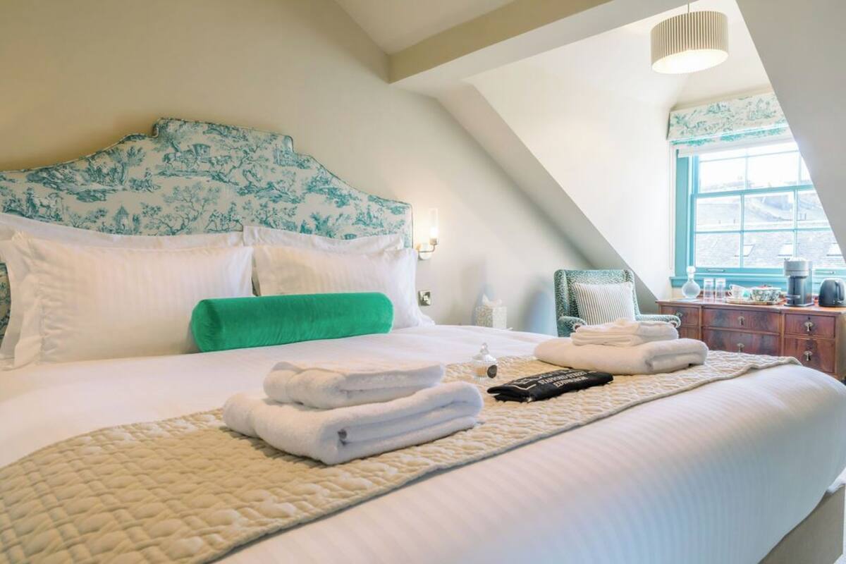 Een net opgemaakt bed met witte en groene beddengoed, een groen kussen en een gewatteerde deken. Er liggen gevouwen handdoeken op het bed, een afstandsbediening en een klein decoratief object.