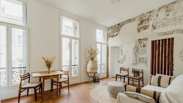 Una habitación con pared de piedra rústica, un banquillo redondo, una mesa de madera con sillas y un jarrón grande con flores secas.