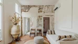 Una habitación con pared de piedra rústica, un jarrón grande con flores, dos sillas de madera, un otoman redondo y una cama con almohadones a rayas.