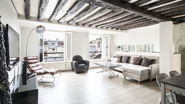 Un salon moderne doté d’un plafond haut, de poutres apparentes, d’un ensemble de canapés, d’une table basse et offrant une vue sur la ville grâce à de grandes fenêtres.