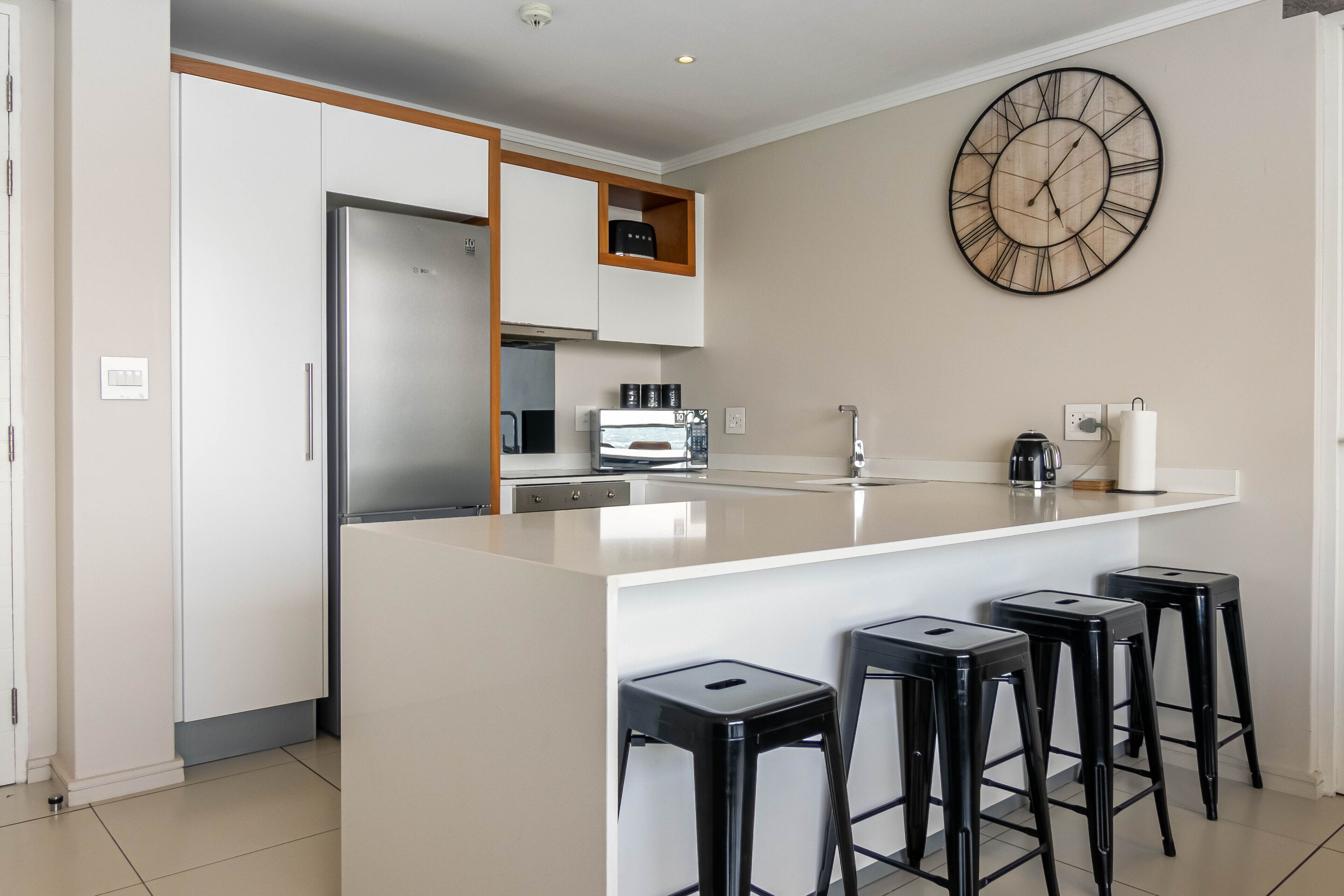 Uma cozinha moderna com ilha central, banquetas, um relógio de parede grande e eletrodomésticos embutidos.