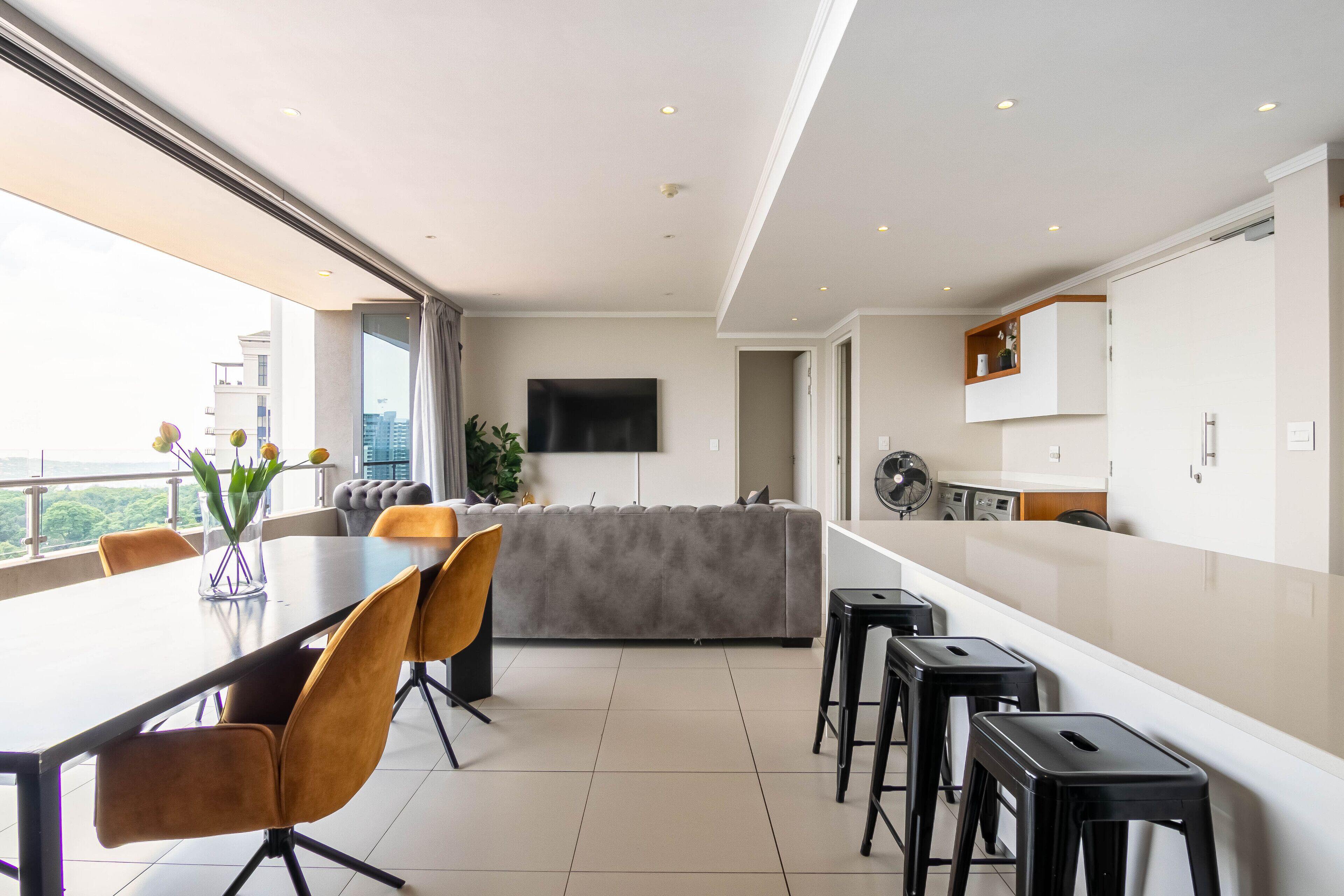 Une cuisine moderne équipée d’un téléviseur à écran plat, d’un coin repas avec une longue table et des chaises, et d’un balcon offrant une vue sur la ville.