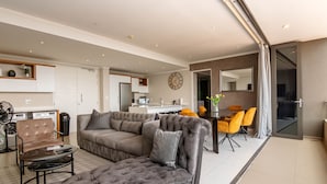 A modern living room with a sectional sofa, a dining area with yellow chairs, a kitchen in the background, and a large mirror on the wall. - Sandton Smart Apartment Seventeen (Sandton)