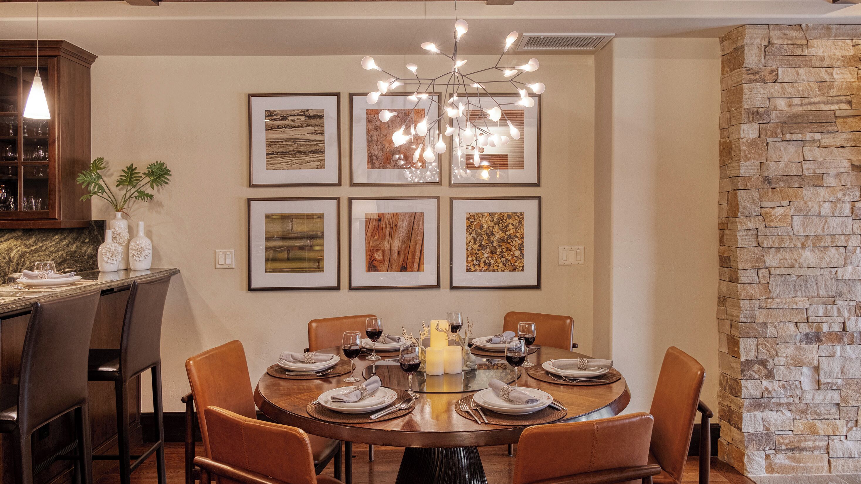 A dining area with a round table set for six, surrounded by six chairs. Above the table, there are six framed artworks on the wall.