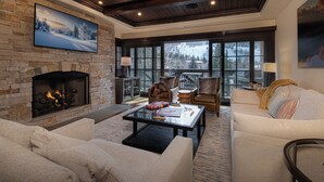 A cozy living room with a stone fireplace, comfortable seating, and large windows offering a view of a snowy landscape. - Walk to Chair 6 + Mountain Views | Chalet Crevasse (Vail)