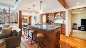 A modern kitchen with a central island, bar stools, wooden cabinetry, and a large window offering a view of trees. - Vail Village Charm + Walk to Lifts| Chalet Schuss (Vail)