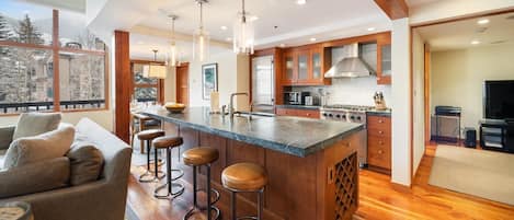 A modern kitchen with a central island, bar stools, wooden cabinetry, and a large window offering a view of trees.