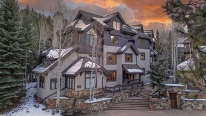A multi-story house with a stone facade, a staircase leading to the entrance, and snow on the roof and ground. - Walk to Arrow Bahn Express | Icegrass Chalet (Edwards)