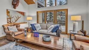 A cozy living room with a leather sofa, wooden coffee table, and a large window with a view of snow-covered trees. - Firepit, Hot Tub + All the Views | Navigator Lodge (Avon)
