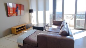 Een moderne woonkamer met een bruine leren bank, een flatscreen-televisie aan de muur, een vloerlamp en een grote schuifglazen deur die leidt naar een balkon met uitzicht op de stad.