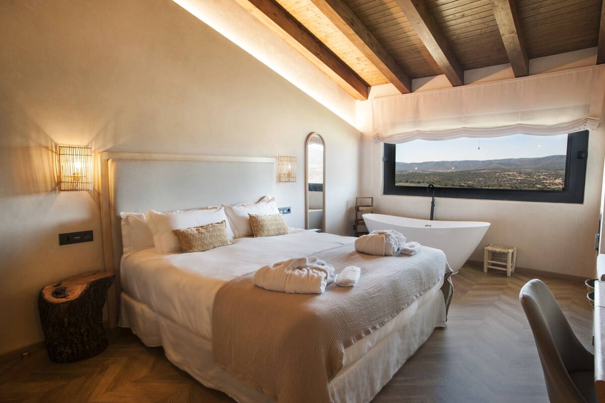 A bedroom with a large bed, a bathtub, and a view of the landscape.