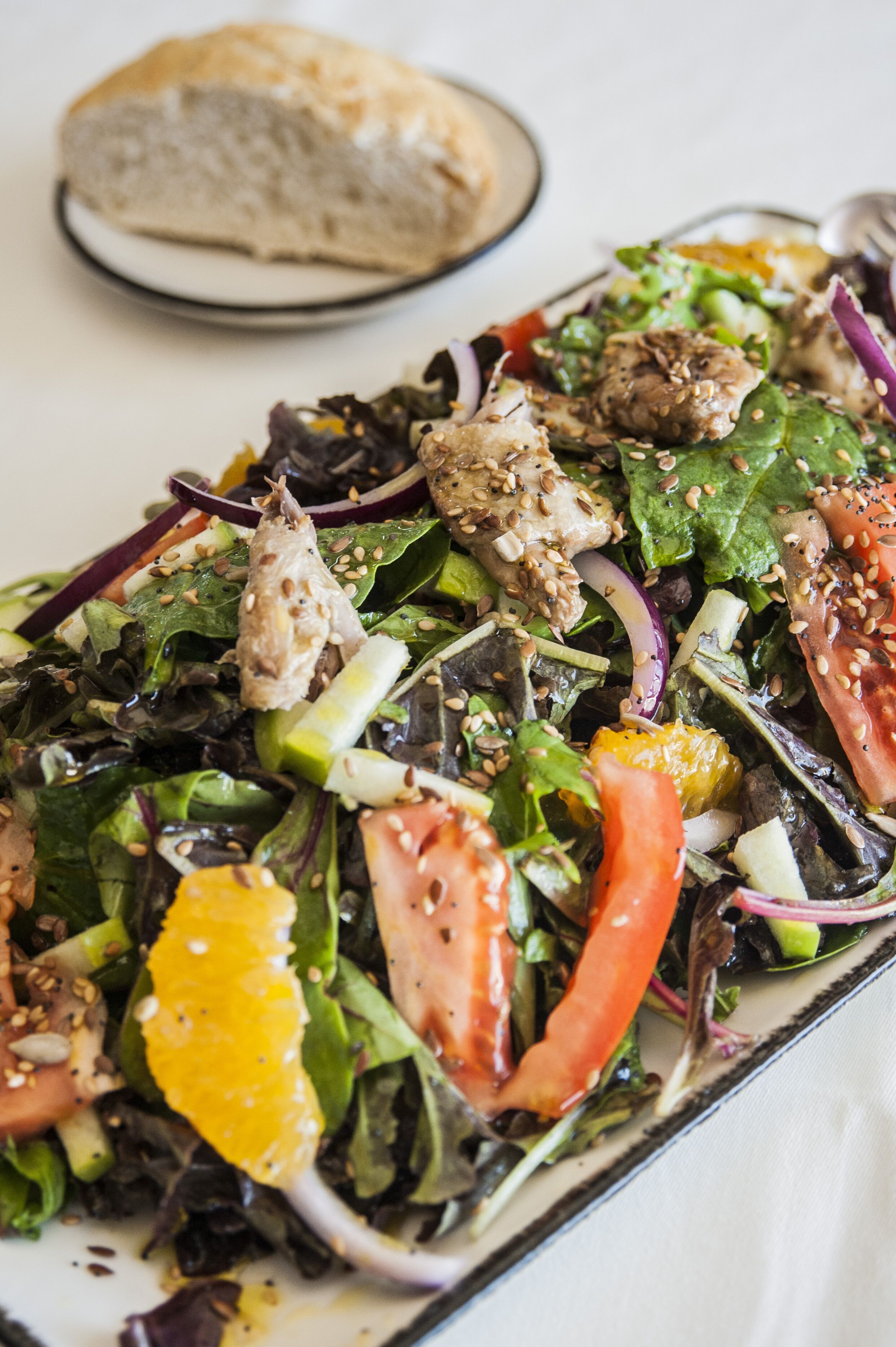 Ensalada de hojas verdes mixtas con diversos vegetales y pollo, acompañada de una rebanada de pan.