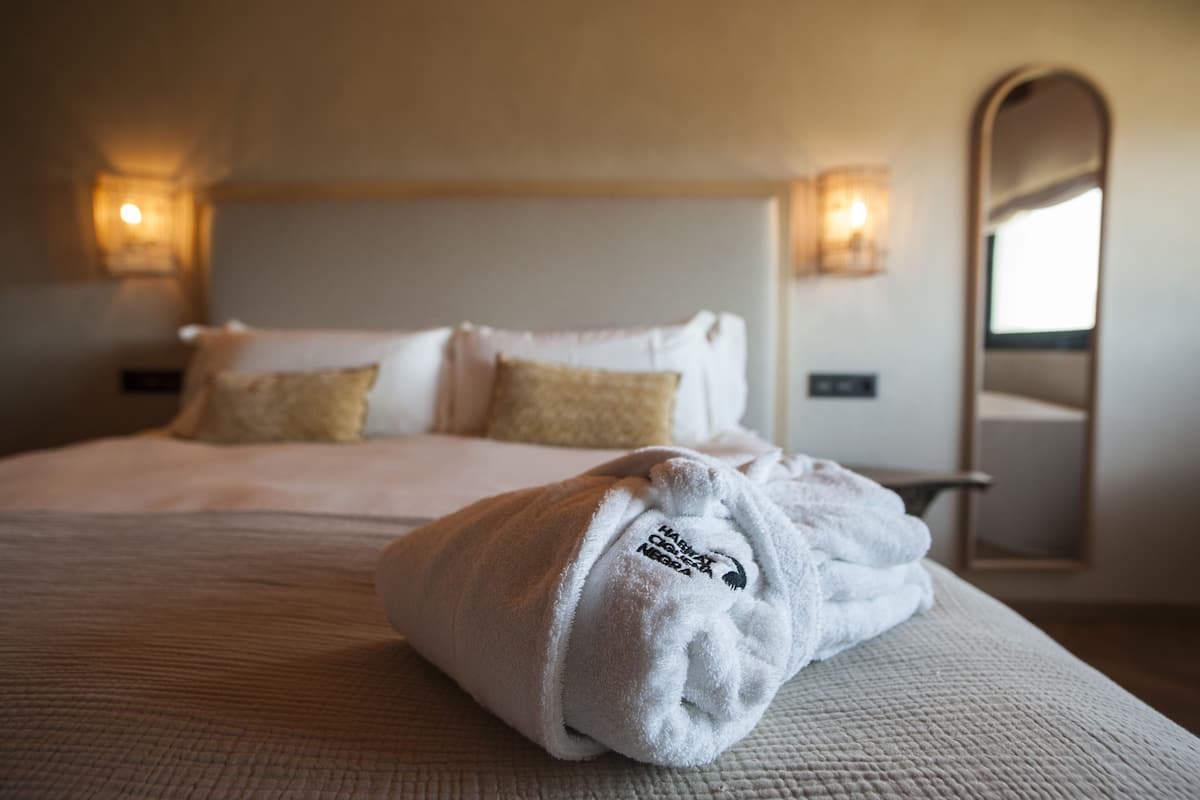 A neatly made bed with a folded towel, two pillows, and a bedside table with a lamp.