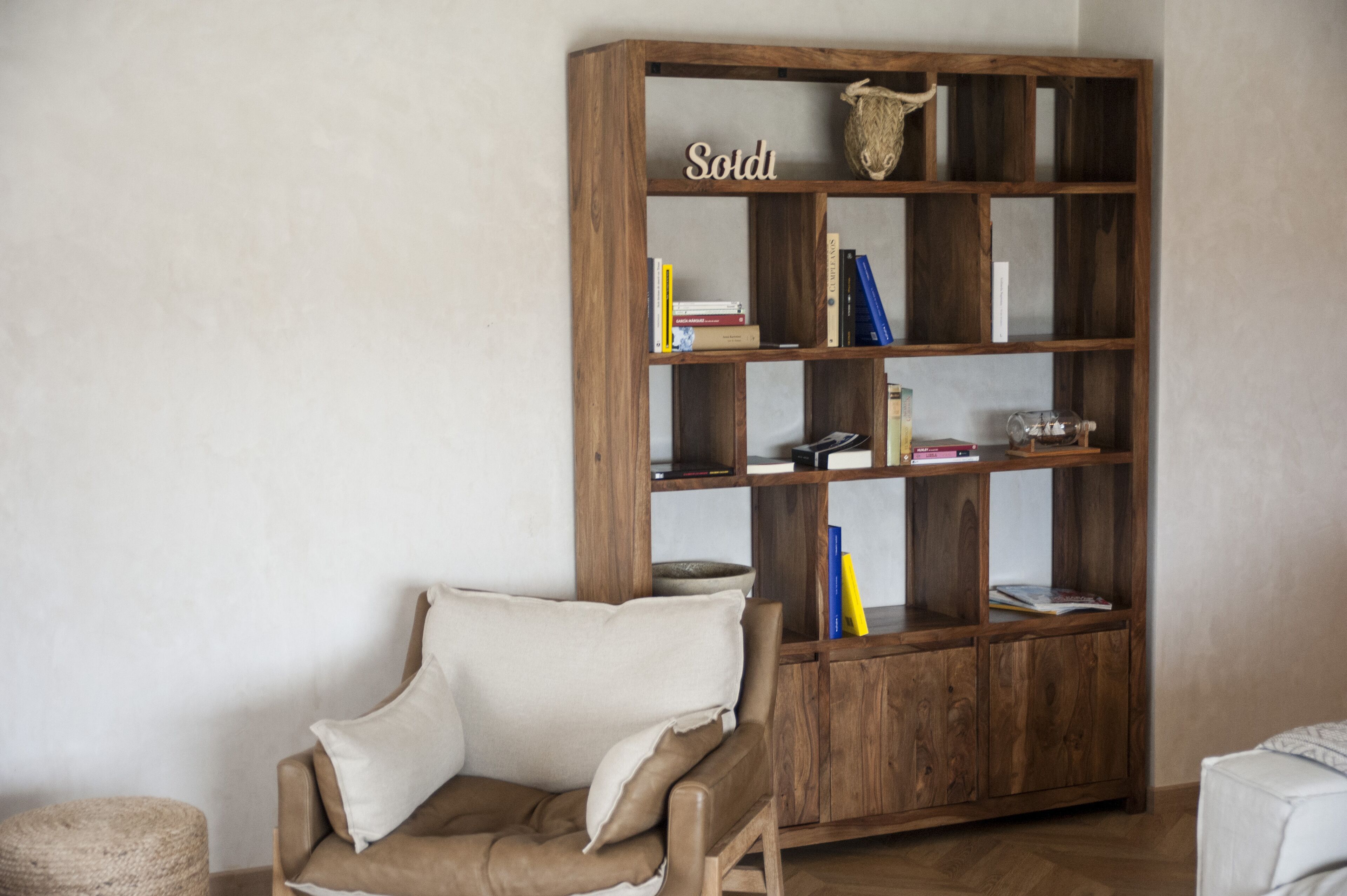Een kamer met een houten boekenkast, een beige fauteuil en een klein rond voetenbankje.