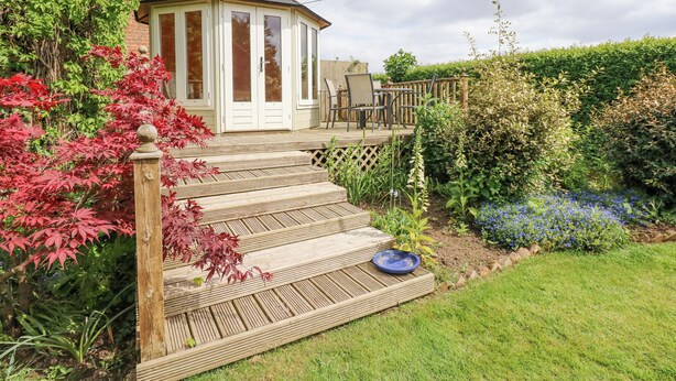 Um deck de madeira leva a uma estufa com porta de vidro, cercada por um jardim com diversas plantas e um canteiro de flores azul.