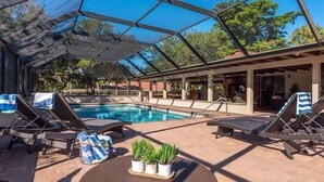 Zona de piscina exterior techada con sillones de descanso y una mesa, rodeada por un recinto de vidrio.