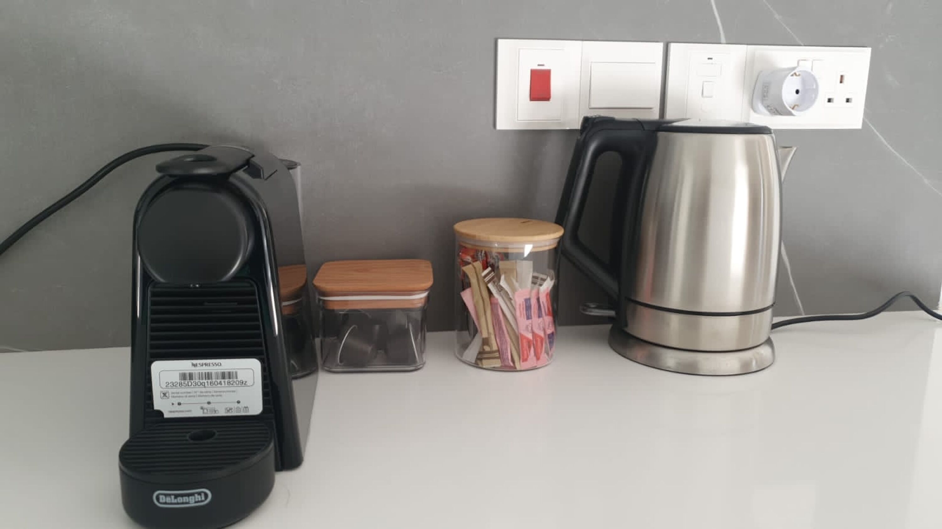 A kitchen counter with a coffee maker, a kettle, a container with papers, and a power outlet.