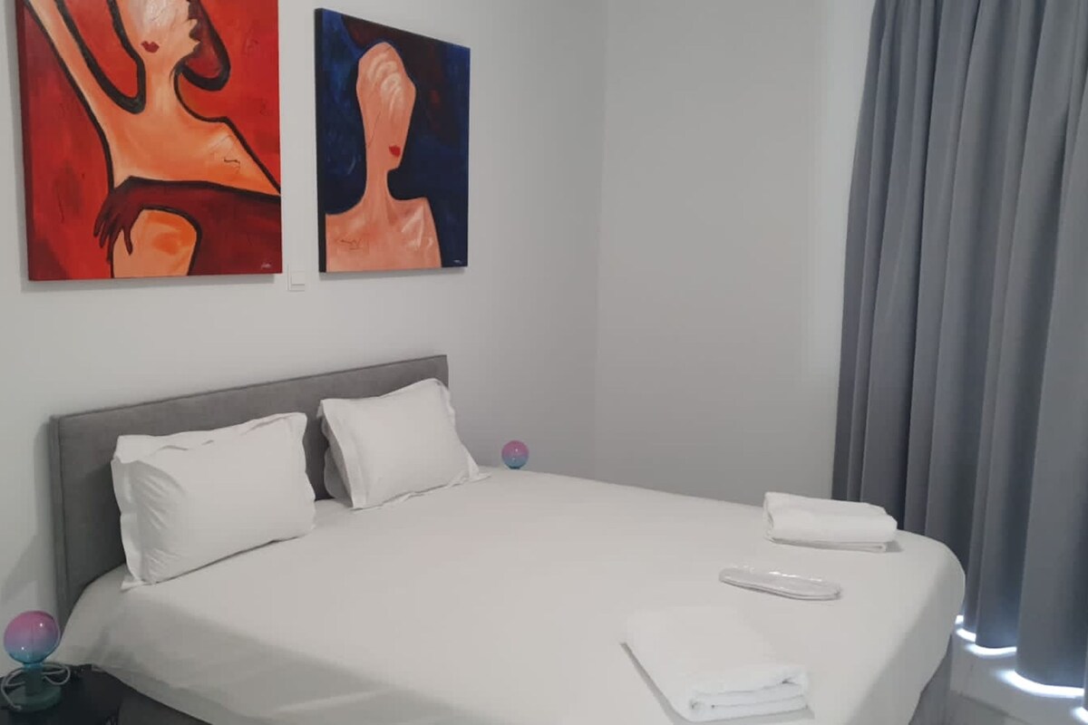 A hotel room with a bed, pillows, a nightstand, and two paintings on the wall.