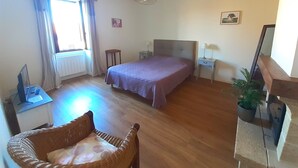 Un dormitorio con cama, sillón, mesita de noche, lámpara, ventana con cortinas y radiador.