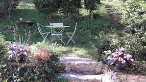 Un jardín con un banco de piedra y una mesa, rodeado de diversas plantas y un sendero.