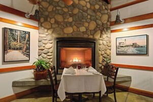 A dining area with a stone fireplace, two framed landscape paintings, and a table set for two.