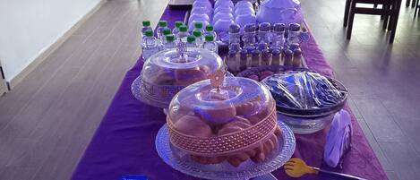 Una mesa de buffet con diversos alimentos y bebidas, cubierta con un mantel morado, ubicada en una sala con ventanas amplias y vistas al exterior.