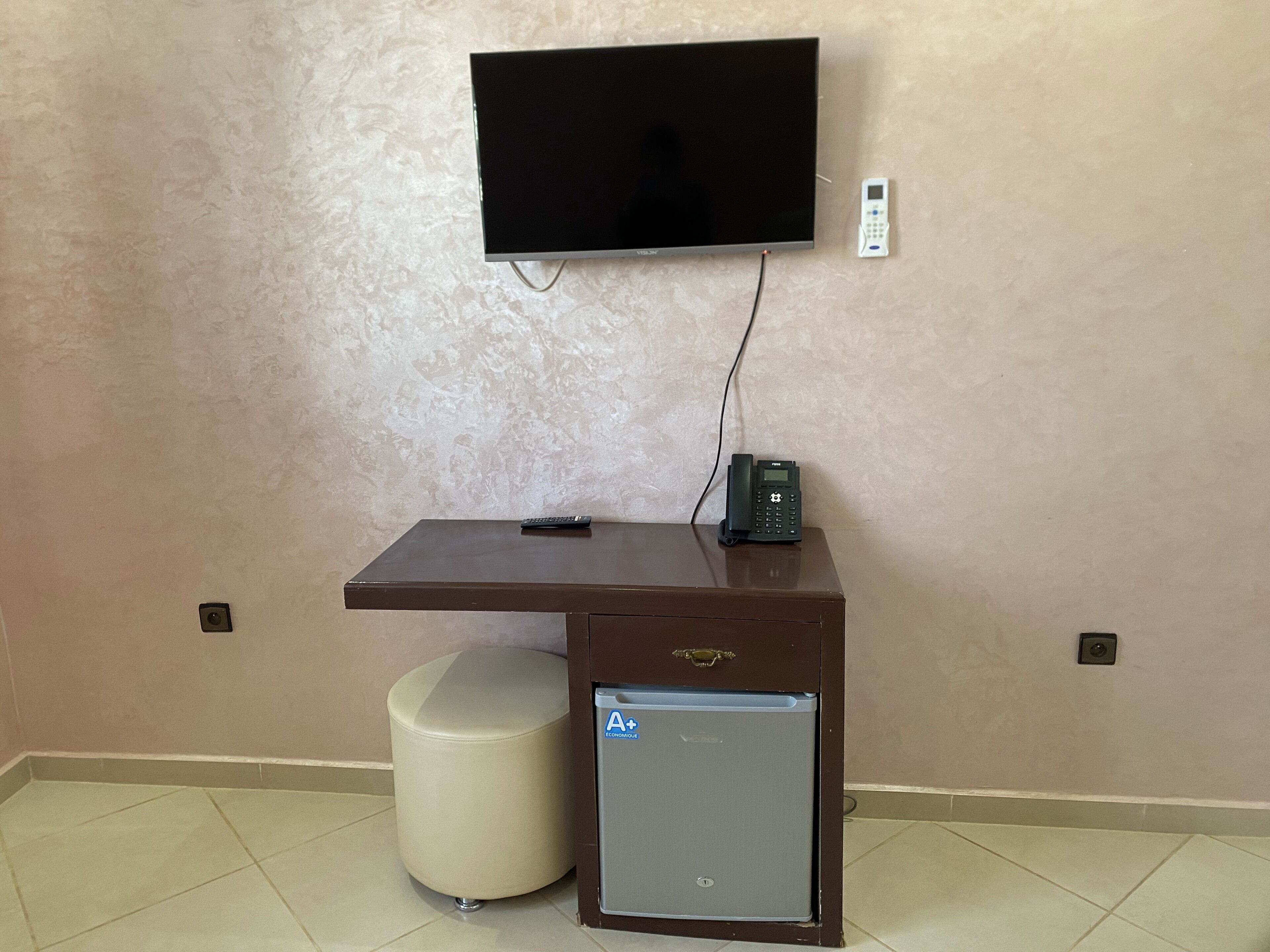 A small, dark wooden desk with a mounted flat-screen TV, a telephone, and a small refrigerator.