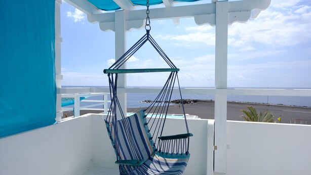 A hammock chair with a striped cushion hanging from a white structure with a blue canopy, overlooking a beach.