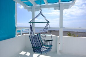 Una poltrona amaca con cuscino a righe sospesa a una struttura bianca con tenda blu, con vista su una spiaggia.
