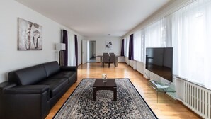 A modern living room with a black sofa, a glass coffee table, and a TV on a glass stand. There is a dining table with chairs in the background, and a large window with curtains. - Nice and big 1 BR flat city center/near lake (D8) (Zürich)