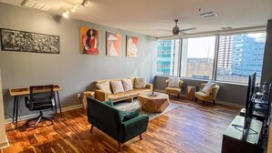 Een ruime woonkamer met houten vloeren, een bank, een salontafel en een tv-meubel. Grote ramen bieden uitzicht op de stad.