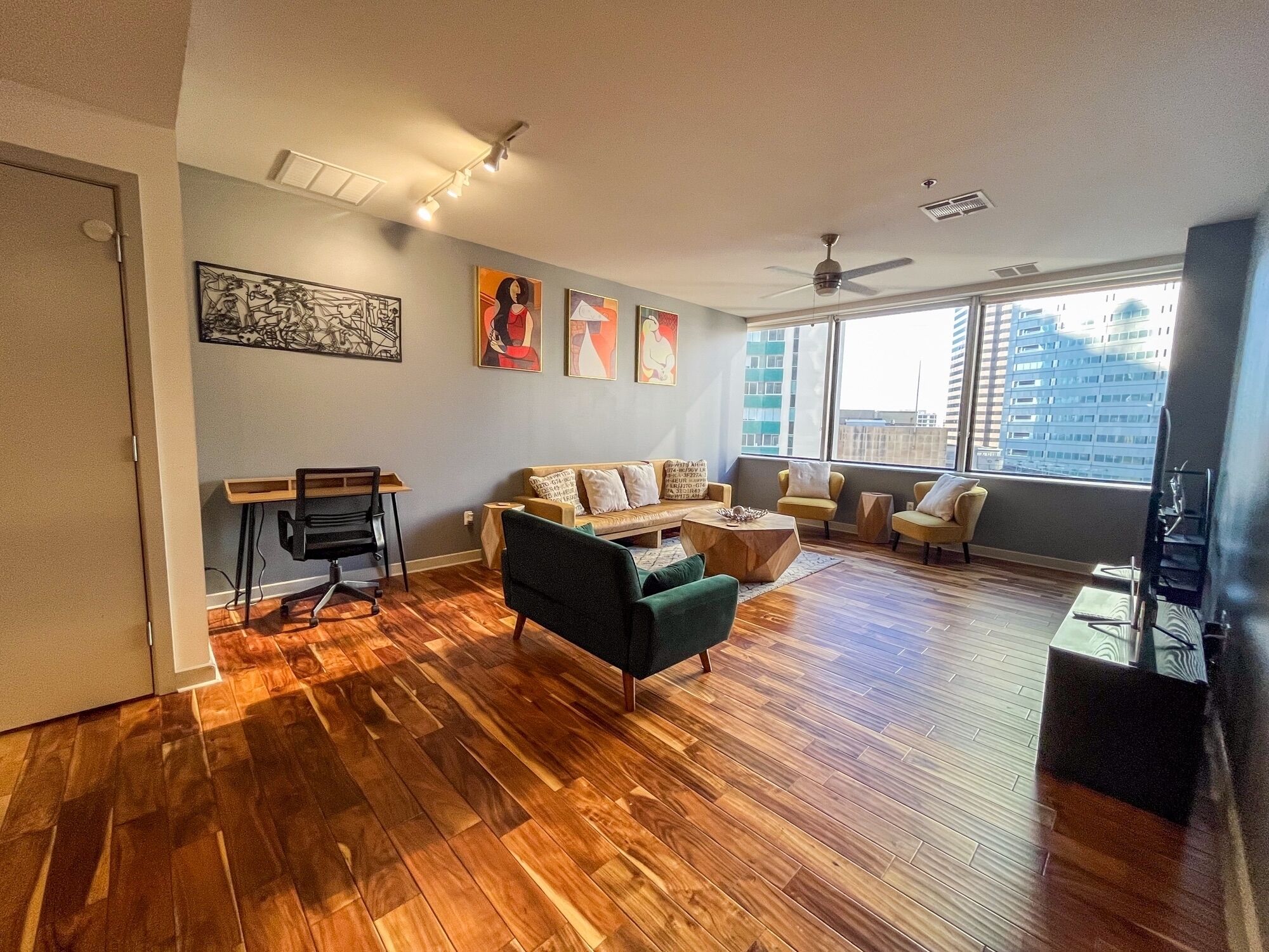 A spacious living room with hardwood flooring, a green armchair, a sofa, and a coffee table. There are several paintings on the wall, a ceiling fan, and a large window offering a view of buildings.
