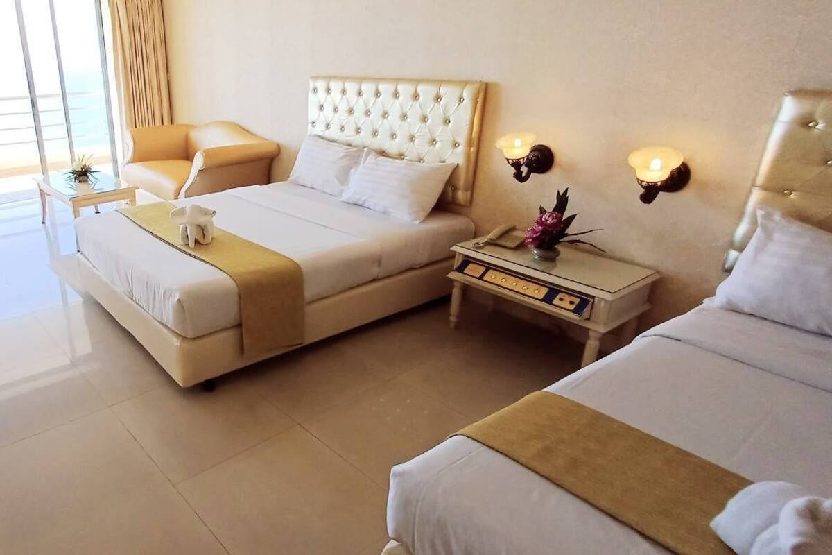 Habitación de hotel con una cama grande, una cama más pequeña, una mesita de noche con lámpara y vistas al océano a través de ventanas amplias.