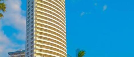 Um hotel alto e cilíndrico com fachada curva, cercado por palmeiras e céu azul límpido.