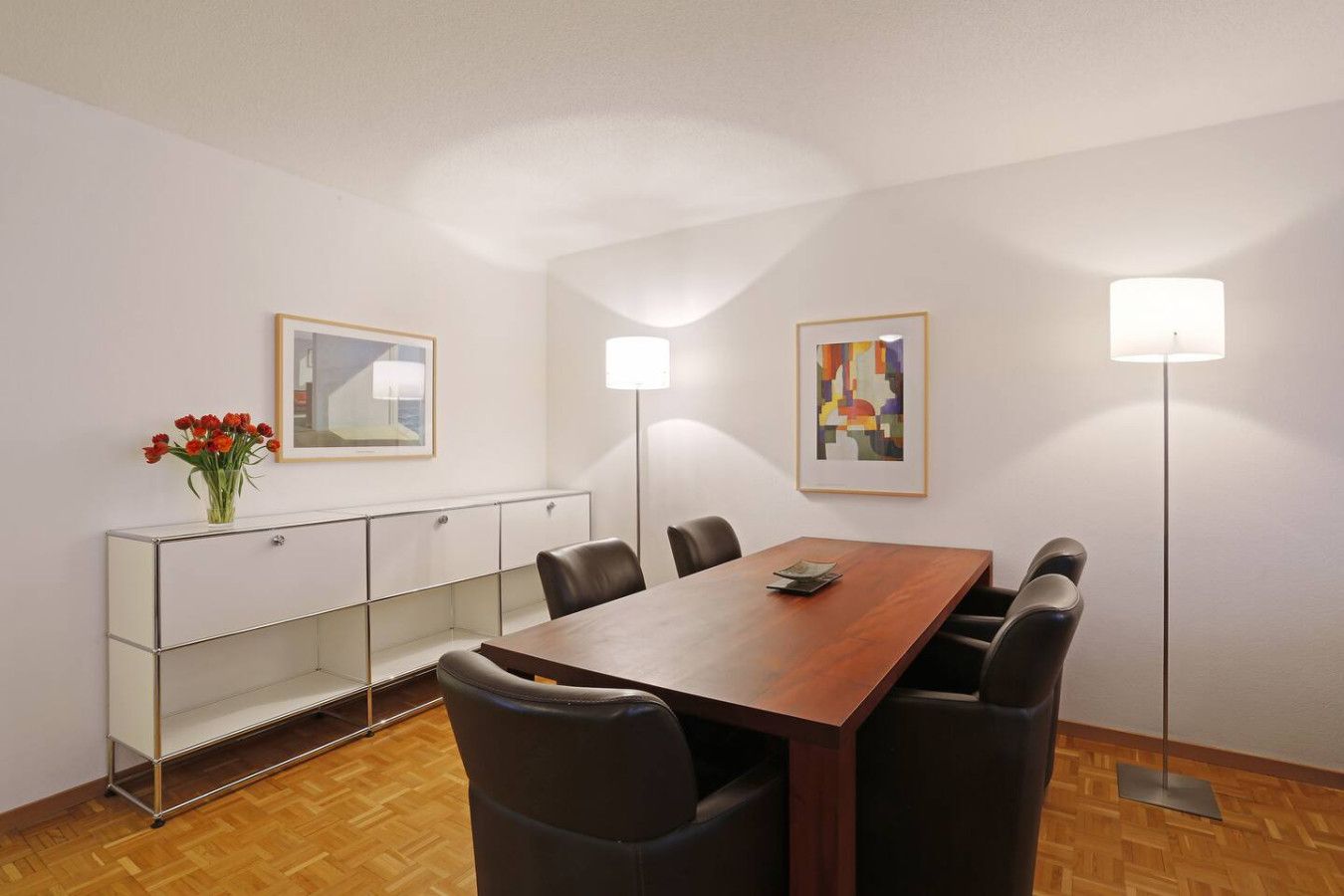 Una sala de conferencias con una mesa de madera, seis sillas, un aparador de vidrio y dos lámparas de pared.