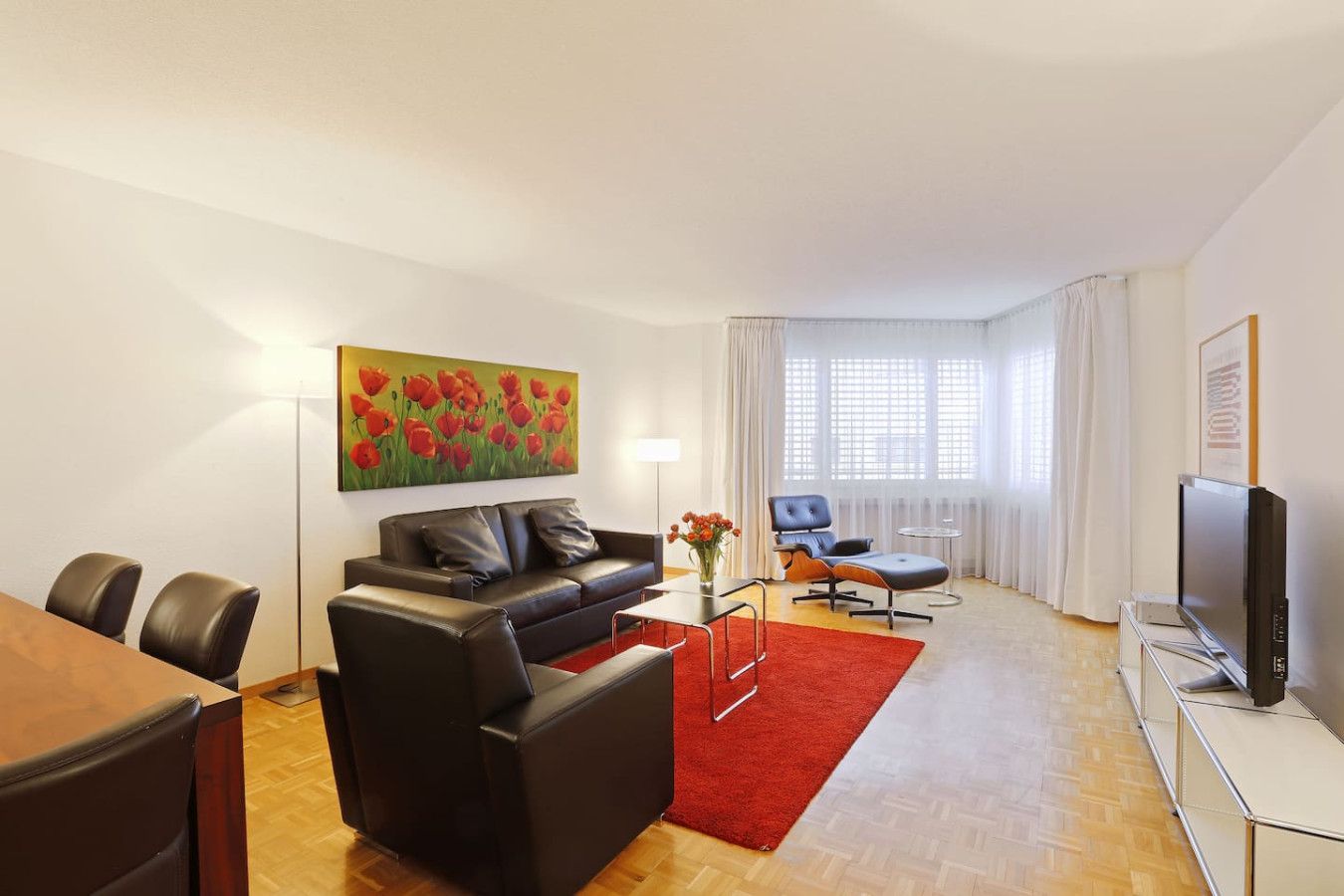 Una sala moderna con un sofá, una mesa de centro y un cuadro de flores rojas. Hay un televisor, una silla y una mesita con un jarrón de flores.