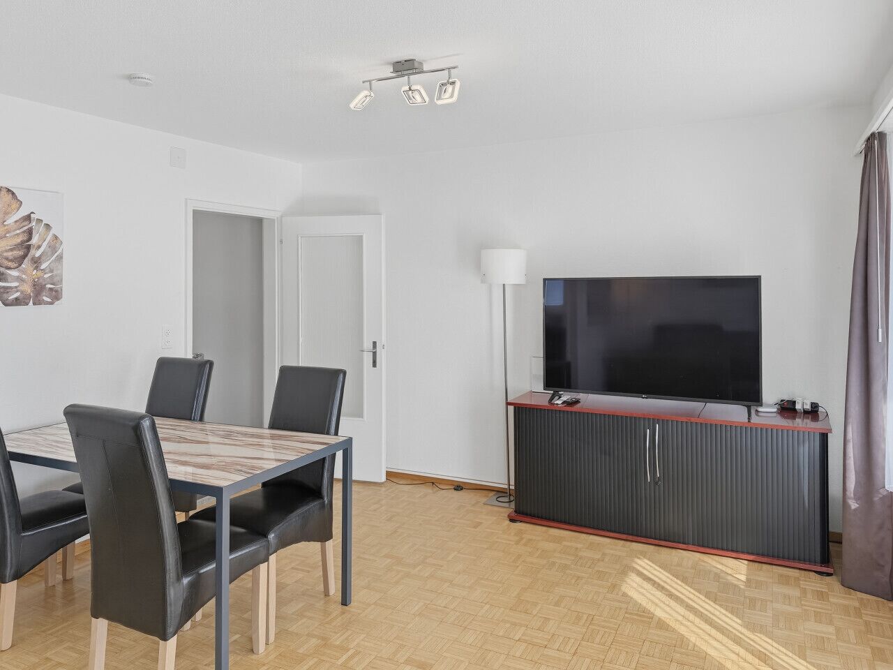 A modern living room with a dining table, chairs, a flat-screen TV, a cabinet, and a lamp.