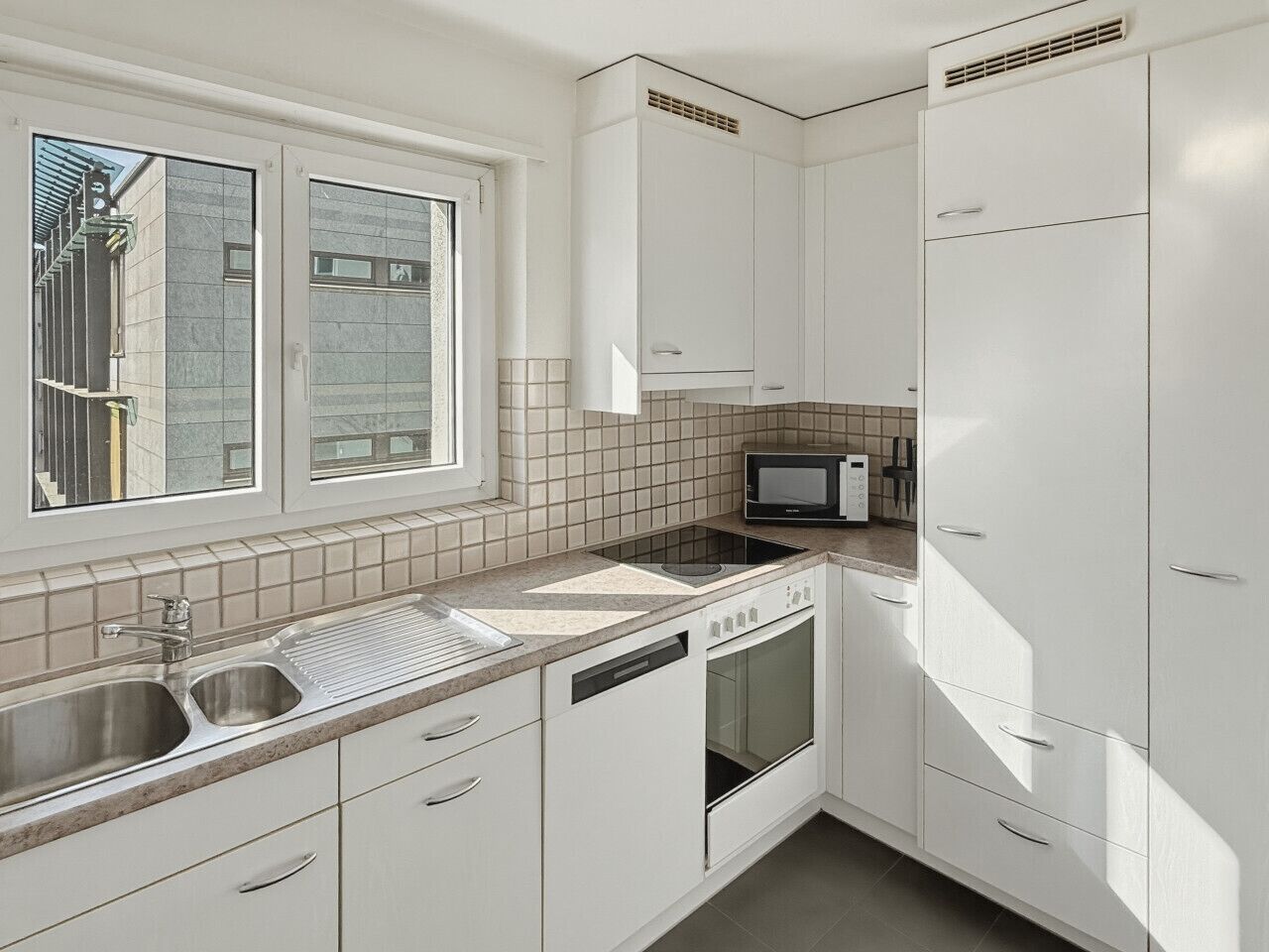 A modern kitchen with a window, stainless steel sink, white cabinets, and a microwave.