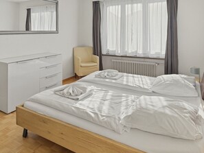 Quarto com cama de madeira, cómoda branca, uma cadeira e janela com cortinas.
