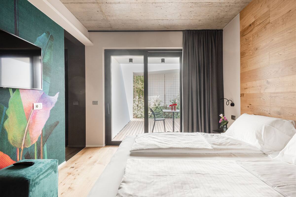 Une chambre à coucher moderne avec un grand lit, une porte-fenêtre coulissante donnant sur un balcon et une peinture abstraite aux couleurs vives accrochée au mur.