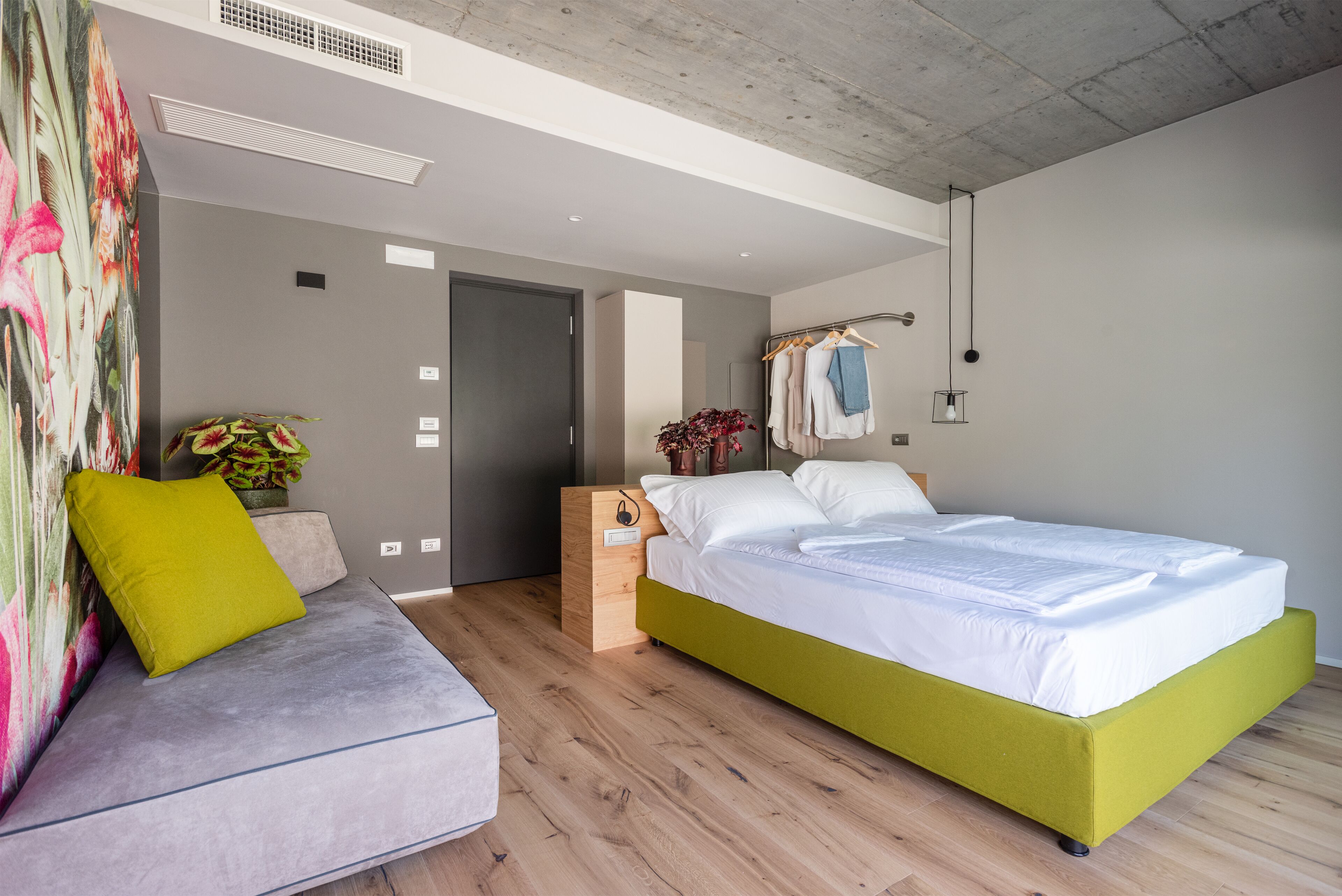 A modern bedroom with a grey sofa, a green bed, wooden flooring, and a concrete ceiling.