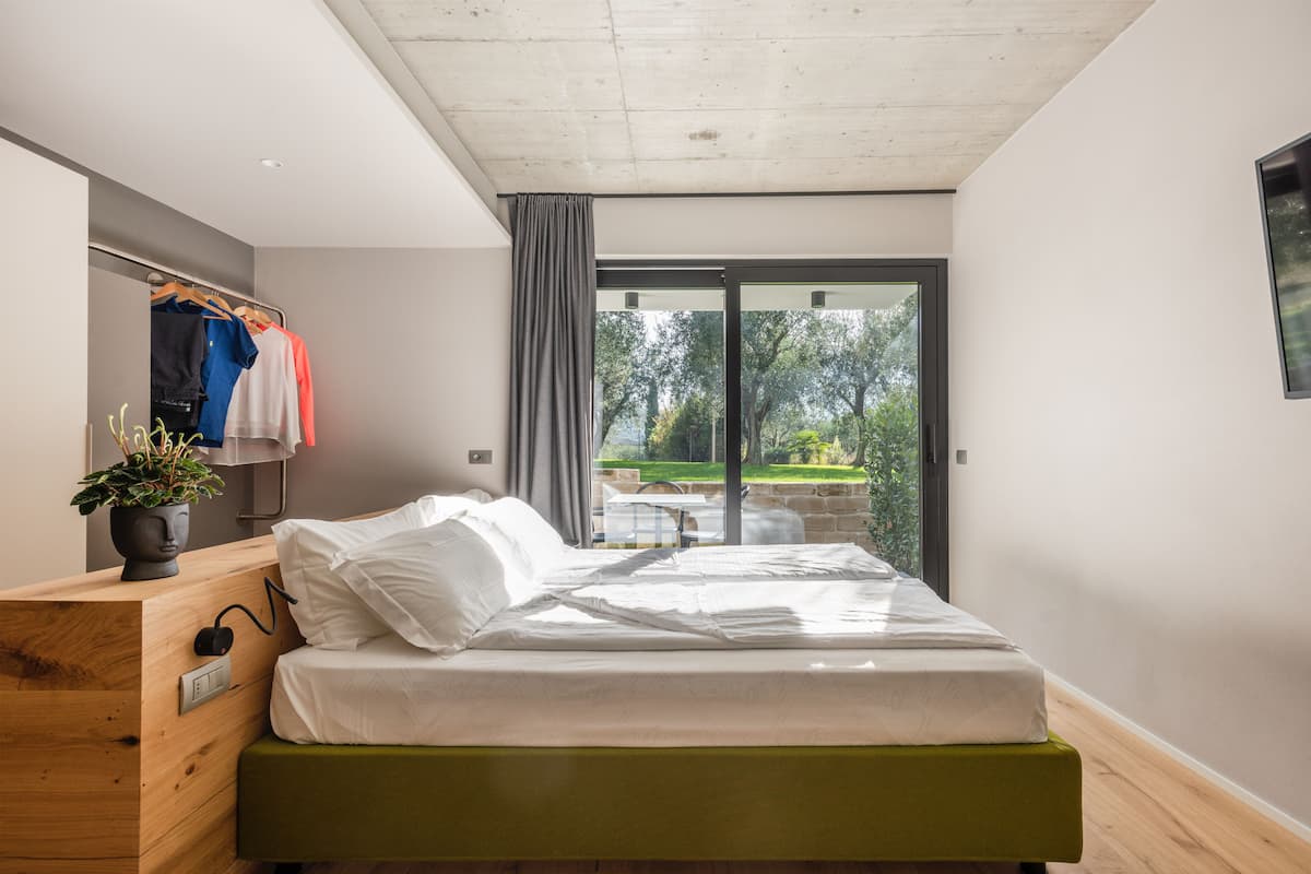 Une chambre à coucher moderne avec un lit, une commode en bois, une plante et une vue sur l’extérieur grâce à une porte-fenêtre coulissante.