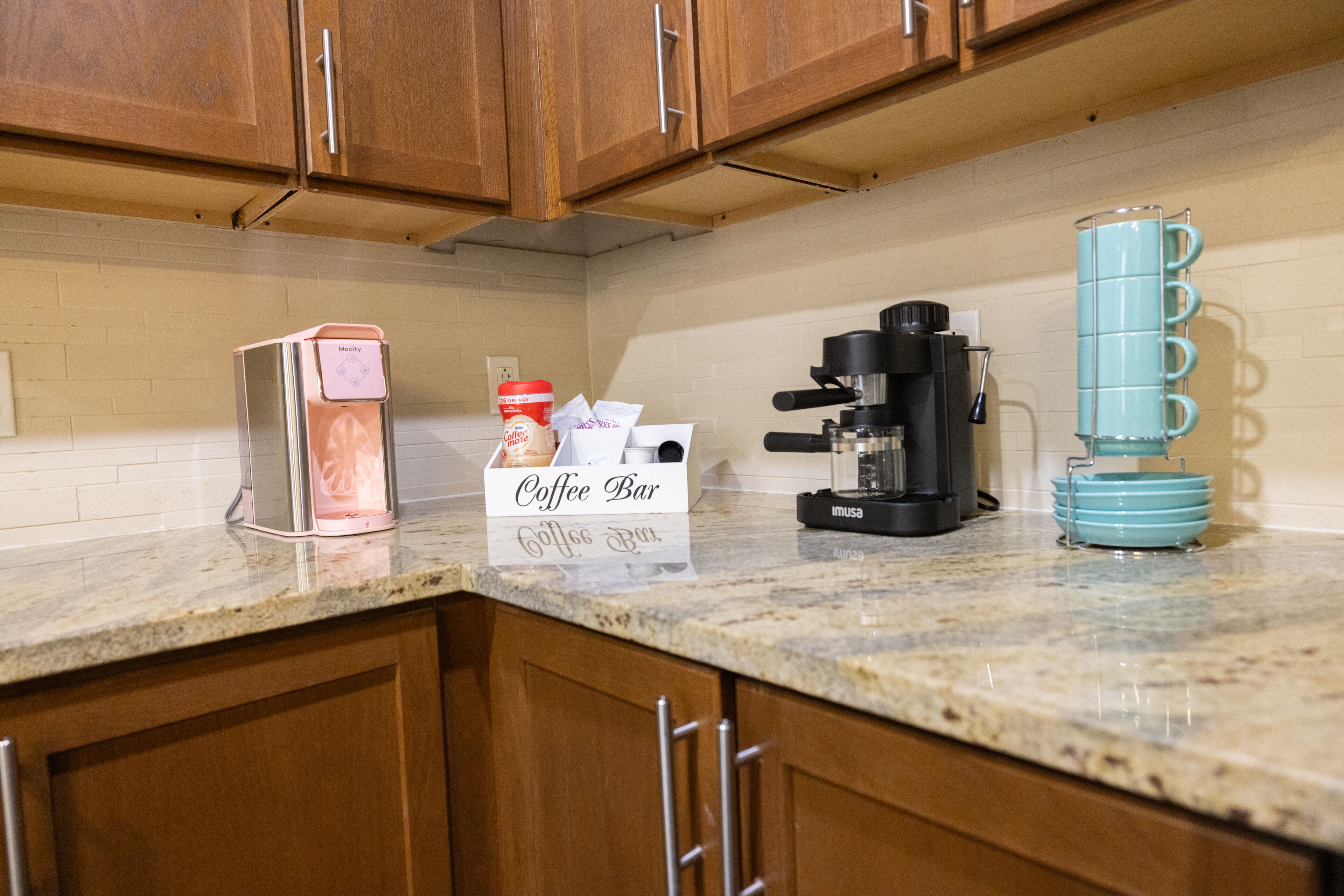 A kitchen with wooden cabinets, a coffee maker, a coffee bar sign, and a stack of blue mugs.