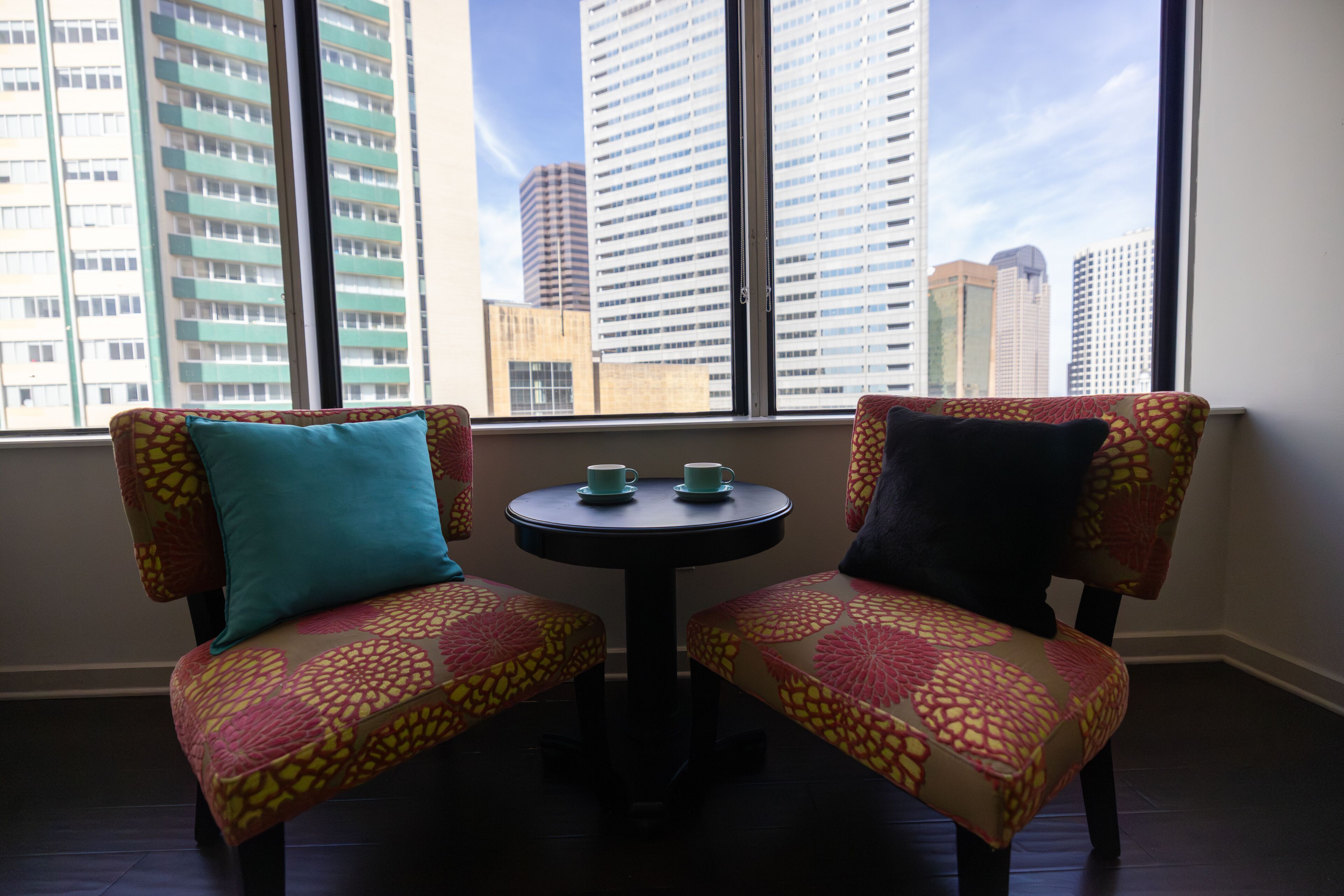 Two chairs with patterned upholstery and a small table with two cups, positioned in front of a window with a cityscape view.