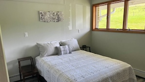 Une chambre à coucher comprenant un lit, deux tables de chevet, une fenêtre donnant sur un paysage verdoyant et un mur orné d’une œuvre d’art florale.