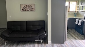 Un salon moderne avec un canapé en cuir noir, un élément décoratif mural et un coin cuisine équipé d’un four à micro-ondes et de placards.