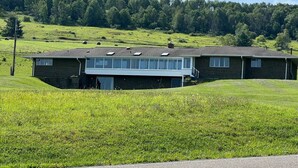 A two-story building with a flat roof, large windows, and a grassy field in the foreground. - Charming Queen Suite w/ Shared Firepit + Game Room (Wellsboro)